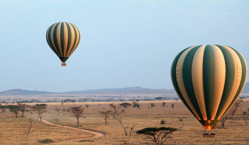 Hot air balloon floating over the Serengeti plains at sunrise with wildlife visible below.